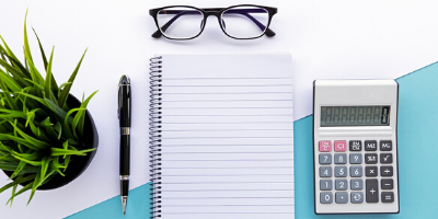 A desk displaying a calculator, glasses a notepad with pen and a small houseplant. 
