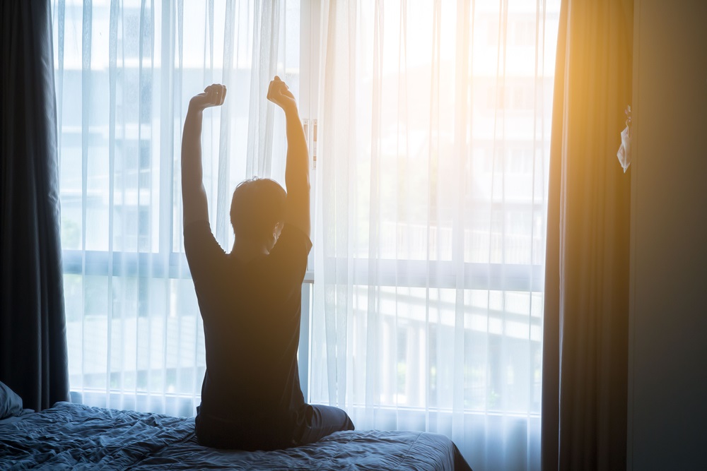 Photo of a man stretching in front of a window with the sun rising outside, as if waking up in the morning. 