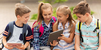 A group of kids sitting outside with backpacks on while browsing the internet on tablets. 