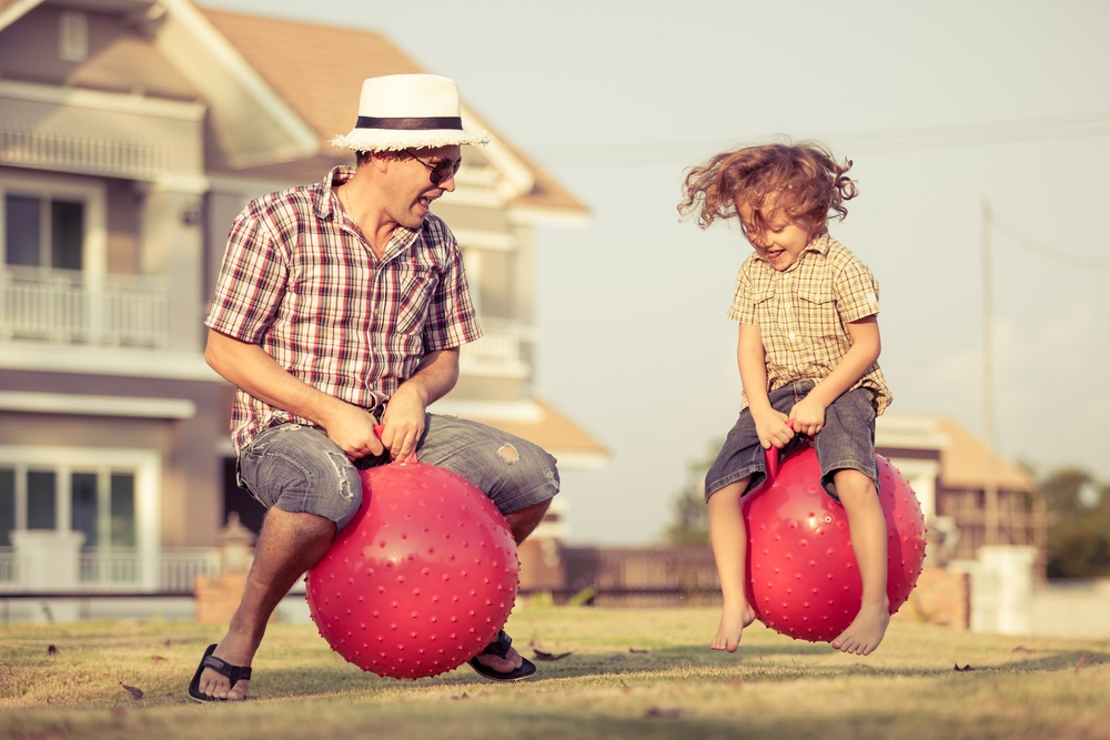 A dad and his son jumping out in the yard on a summer day with red, bouncy balls.