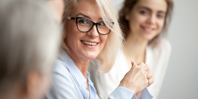 A senior adult woman seated in a work setting while smiling and mentoring younger colleagues. 
