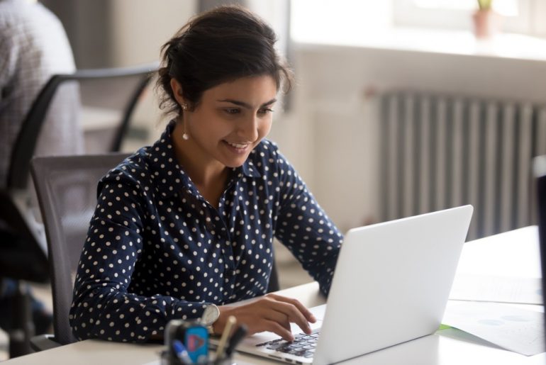 Young Indian professional sitting at a desk, working on a computer and smiling.