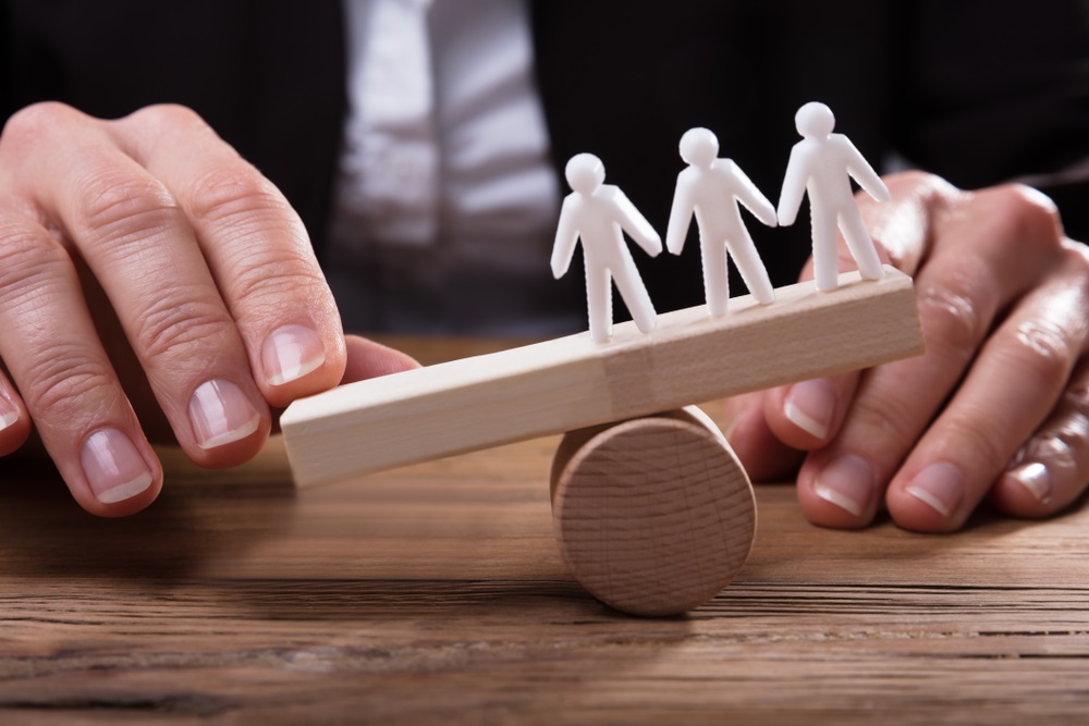 Image of three stick figures on a balance being held up as a hand pushes the other side down.