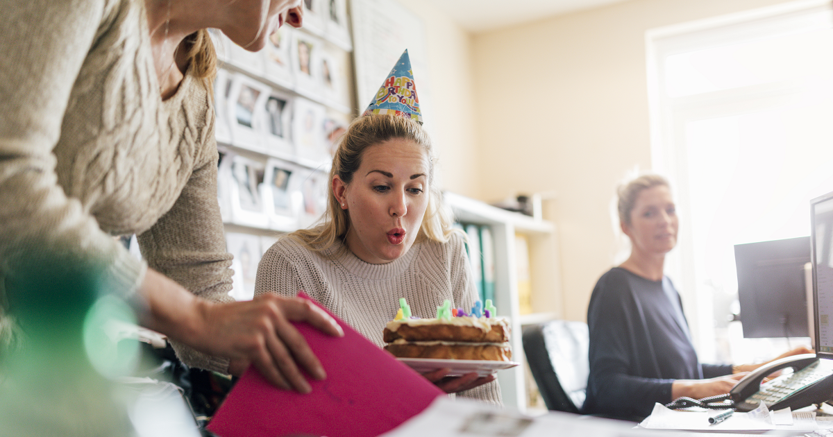 Woman wearing party hat and blowing out candle on birthday cake in office with coworkers