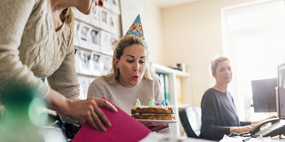 Woman wearing a party hat and blowing out candle on birthday cake in office with coworkers