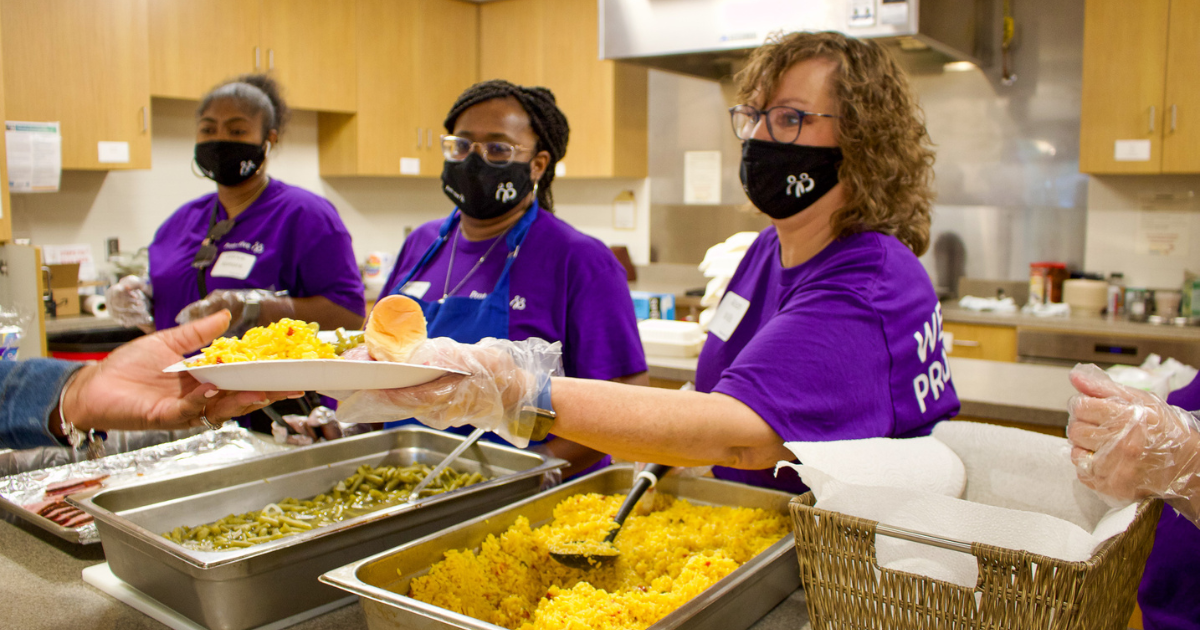Three women in purple shirts handing plates of food across the pickup line