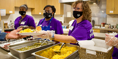 Three women handing a plate of food across a buffet of food.